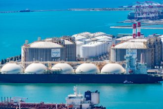 Vista aérea de um terminal de gás natural liquefeito, com tanques de armazenamento e navios-tanque junto à costa.
