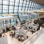 Pessoas circulam e conversam em uma feira de negócios dentro de um moderno pavilhão de exposições, entre estandes de empresas.