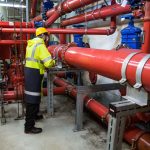 Profissional com equipamento de segurança inspeciona tubulações industriais em uma instalação de processamento de biogás.