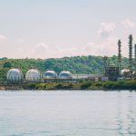 Instalação industrial costeira de armazenamento de gás, com tanques esféricos e chaminés.